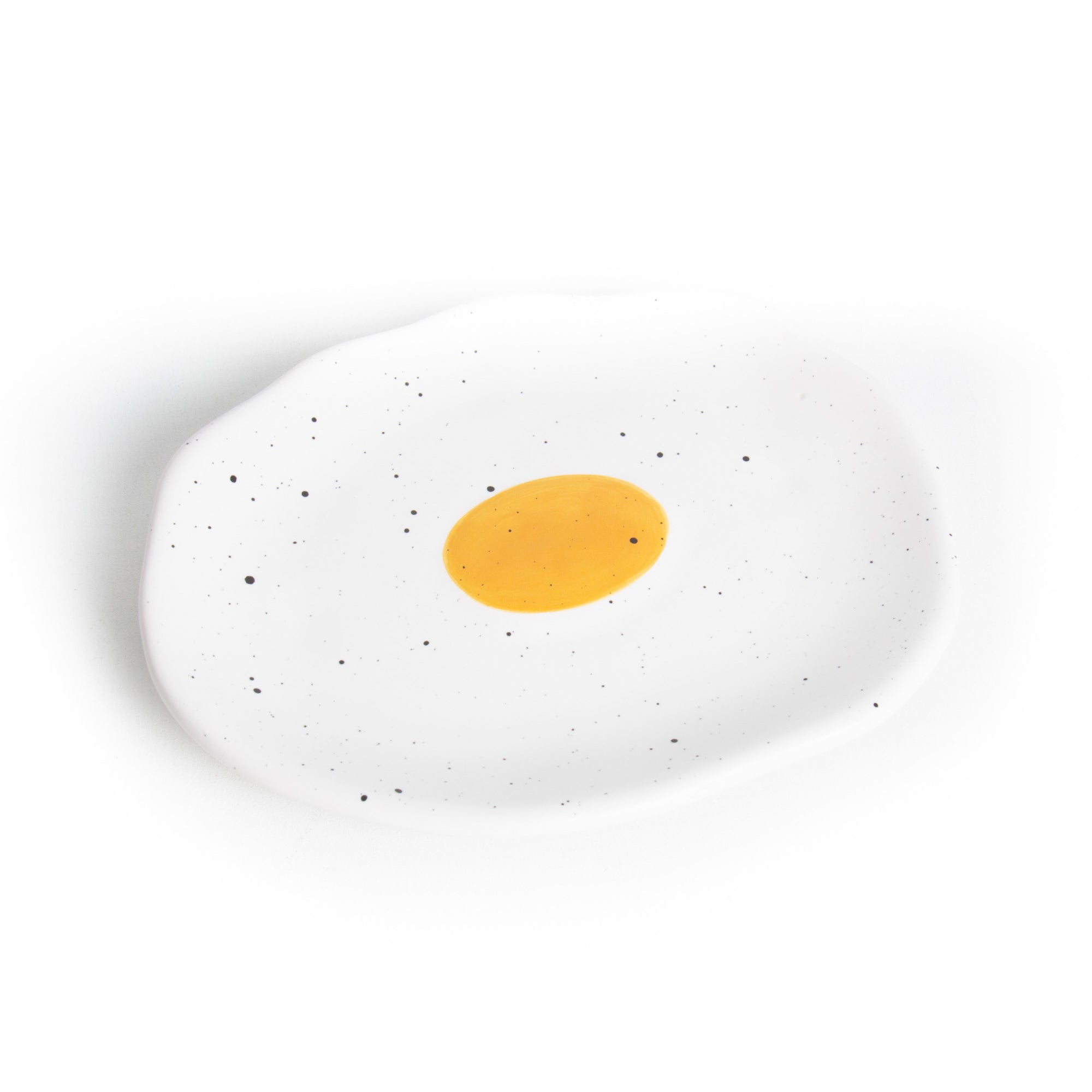 A ceramic jewellery tray designed to resemble a fried egg with a white base and a yellow center, featuring black speckles.