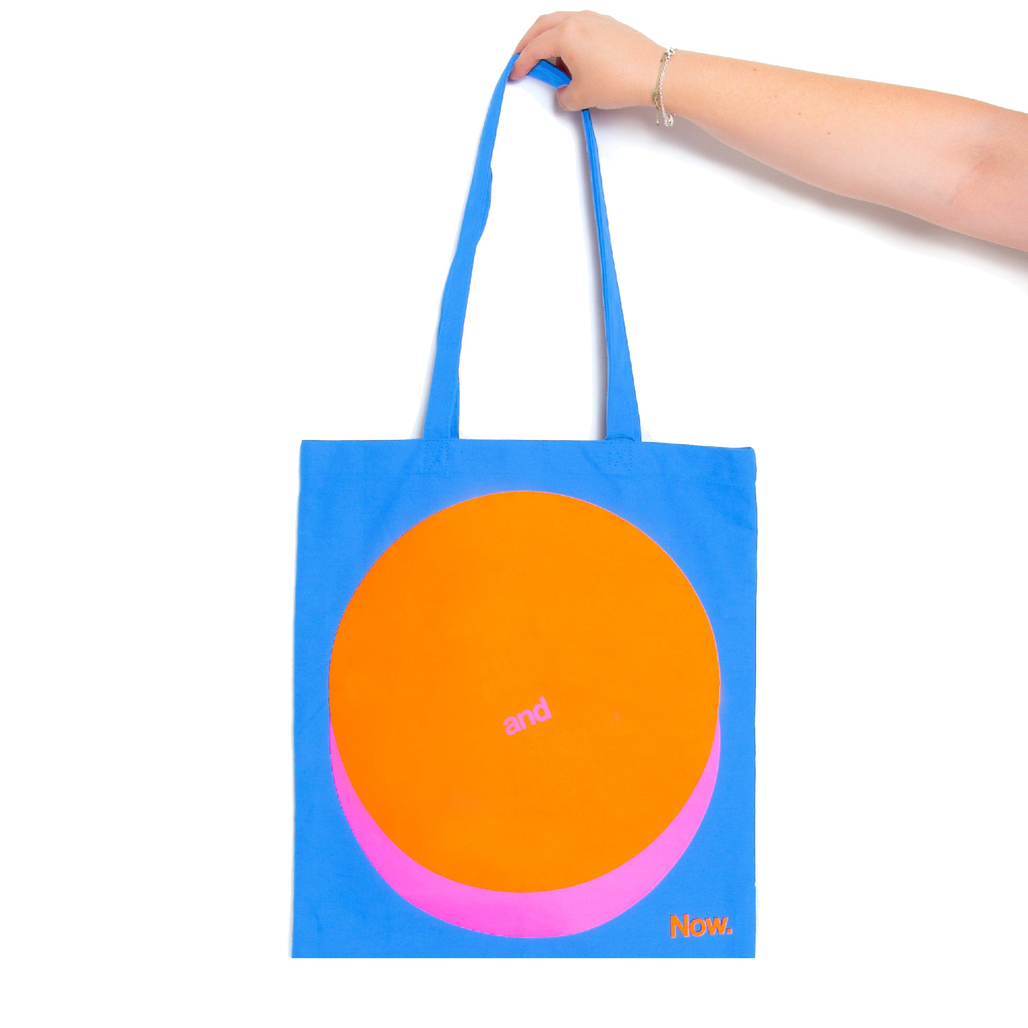 A person holding a blue tote bag with a large orange circle design on the front and the text 'and now' across the orange circle.