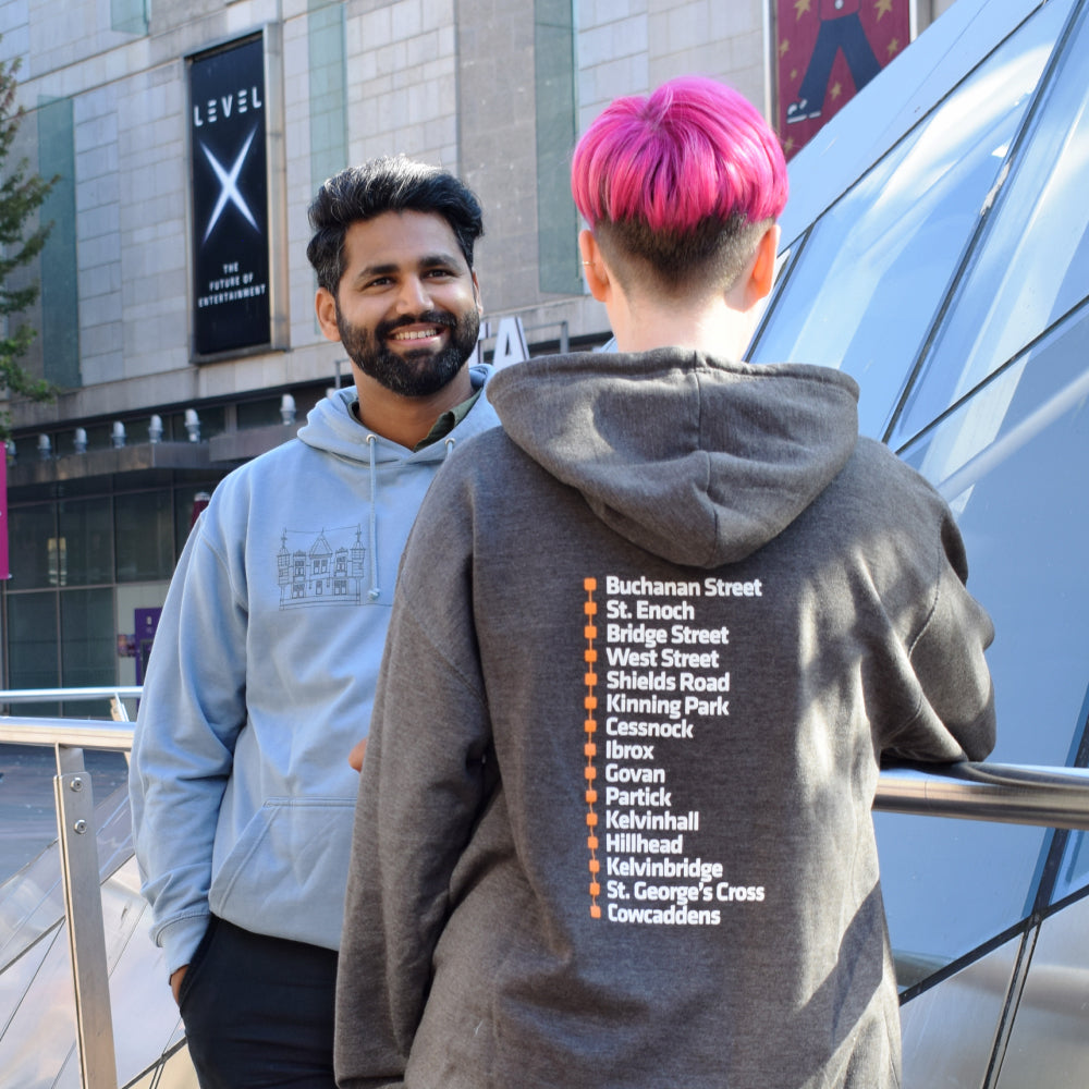 Two people standing outdoors, one wearing a hoodie with Subway station list printed on it.