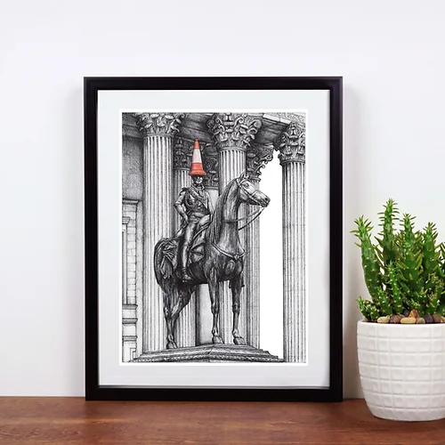 Framed artwork of a classical statue with a traffic cone on its head, placed on a wooden surface next to a plant.